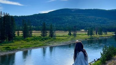 Traveller sitting on old log alongside a stream of water looking towards the mountains in the distance